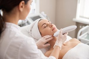 Cosmeticiană aplicând manipulul unui aparat HIFU pe conturul facial al unei cliente